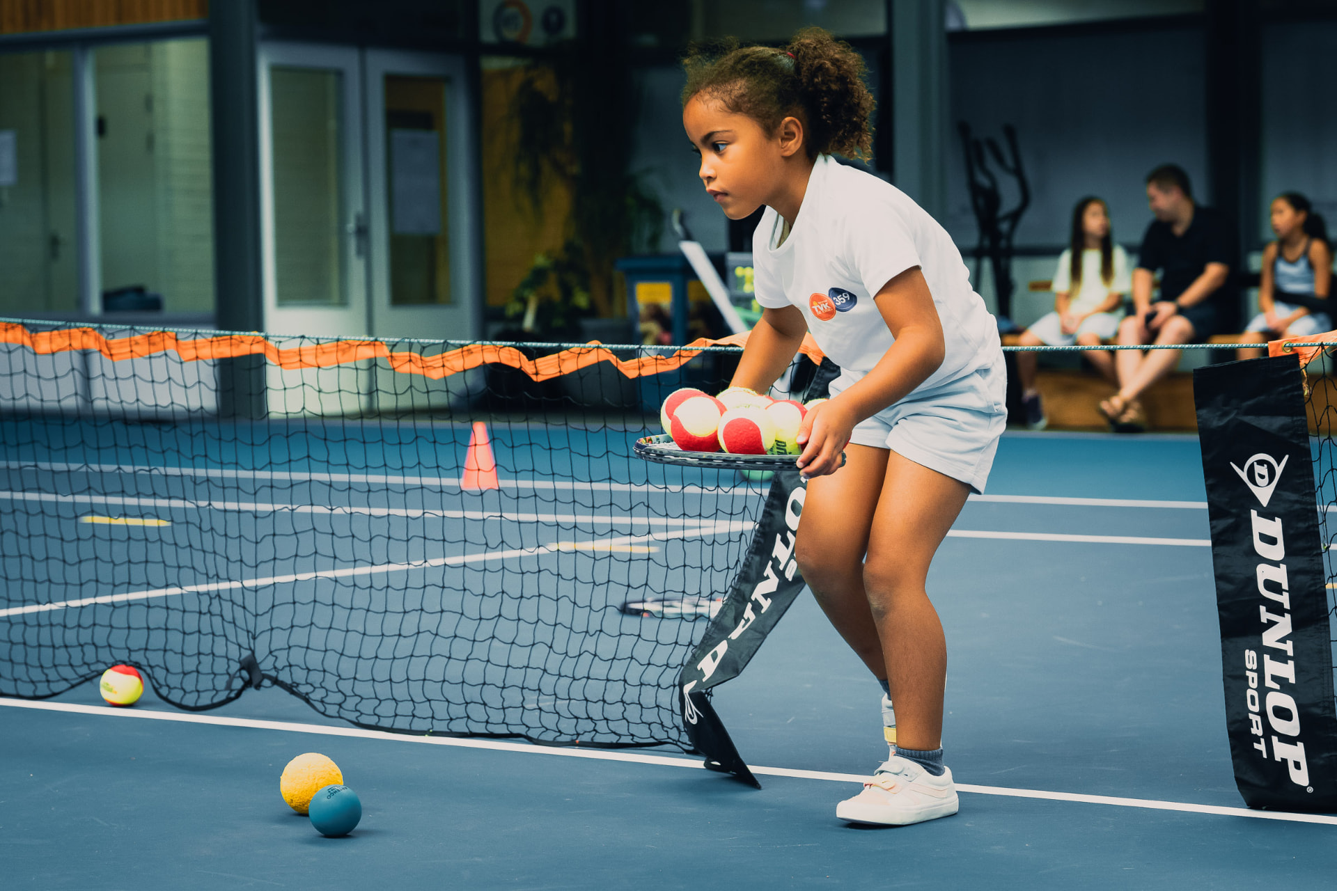 Meisje verzamelt oranje-punt en rode ballen bij het net tijdens tenniskids-les — 359.tennis