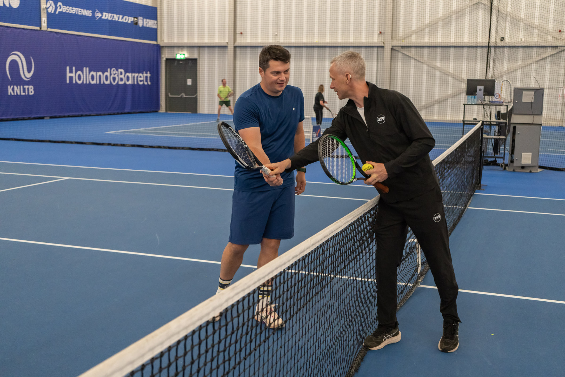Coach guides an adult player at the net on an indoor court at NTC Amstelveen — 359.tennis