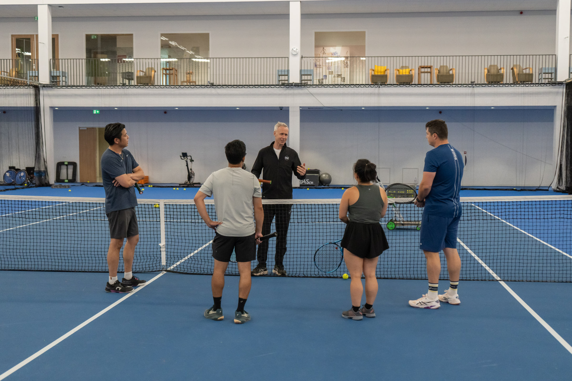 Coach briefs a group of adults at the net on an indoor court at NTC Amstelveen — 359.tennis