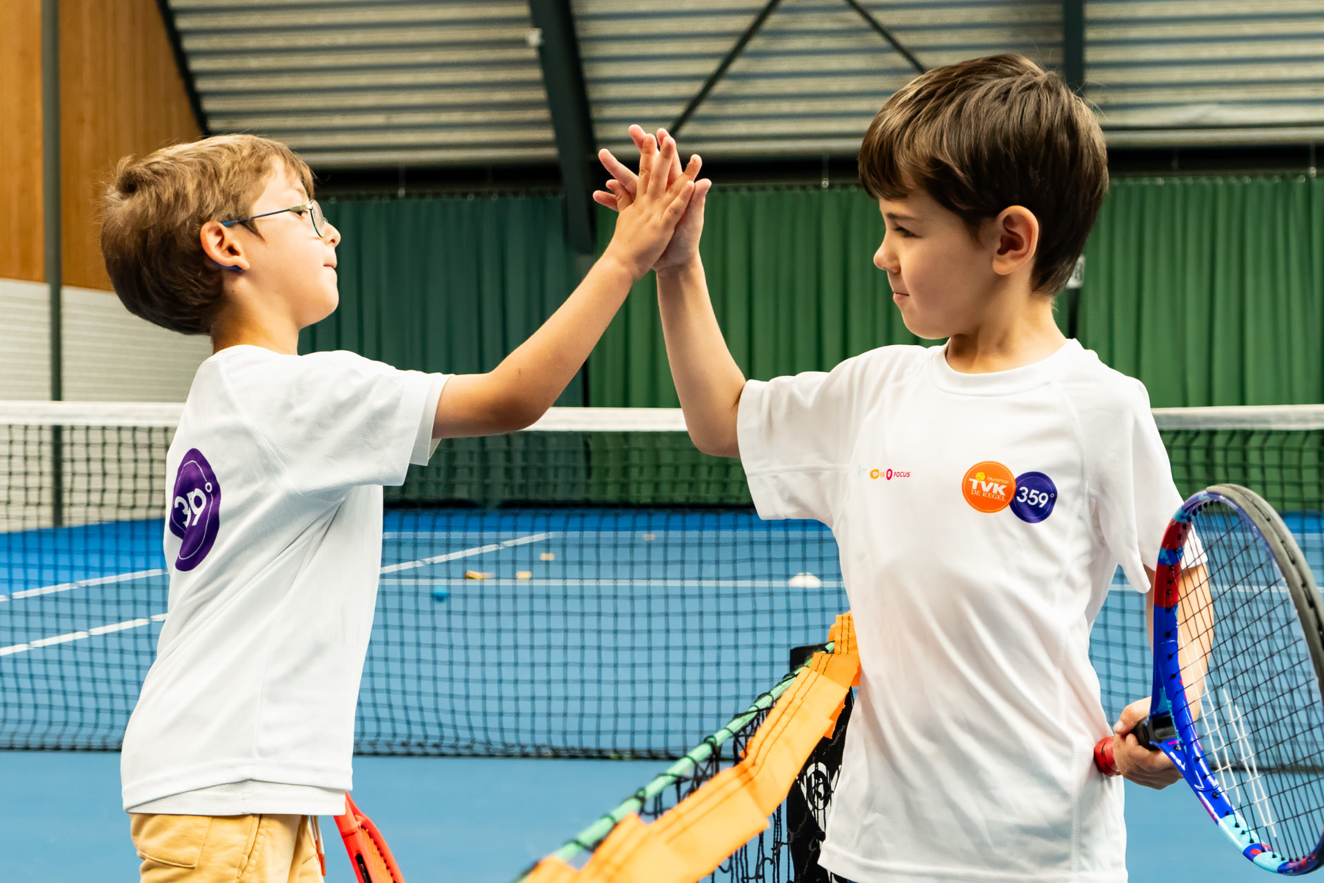 Twee jongens geven elkaar een high five na een punt tijdens indoor tennisles op NTC Amstelveen — 359.tennis