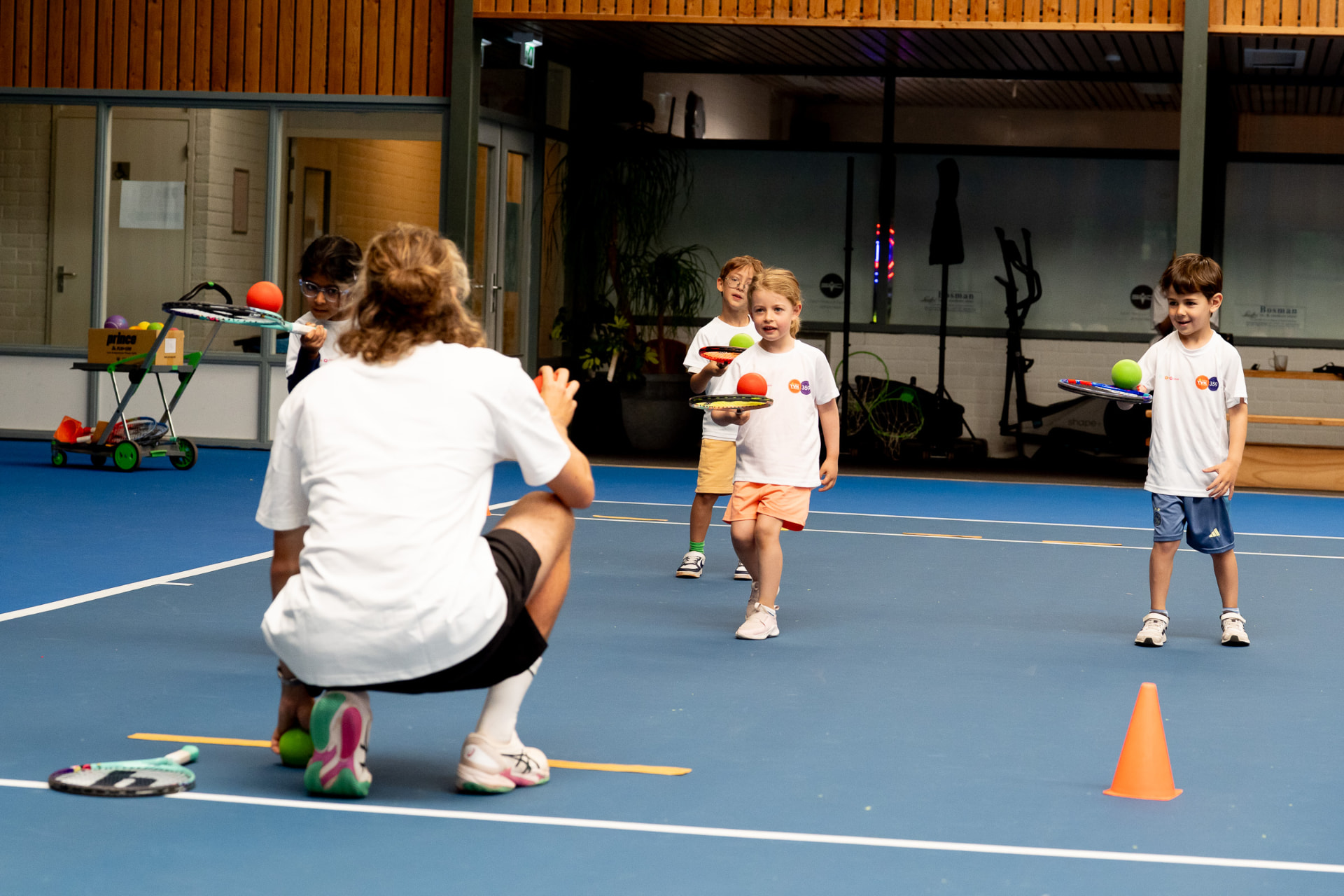 Coach guides young tenniskids through a balance and ball-control drill on an indoor court — 359.tennis