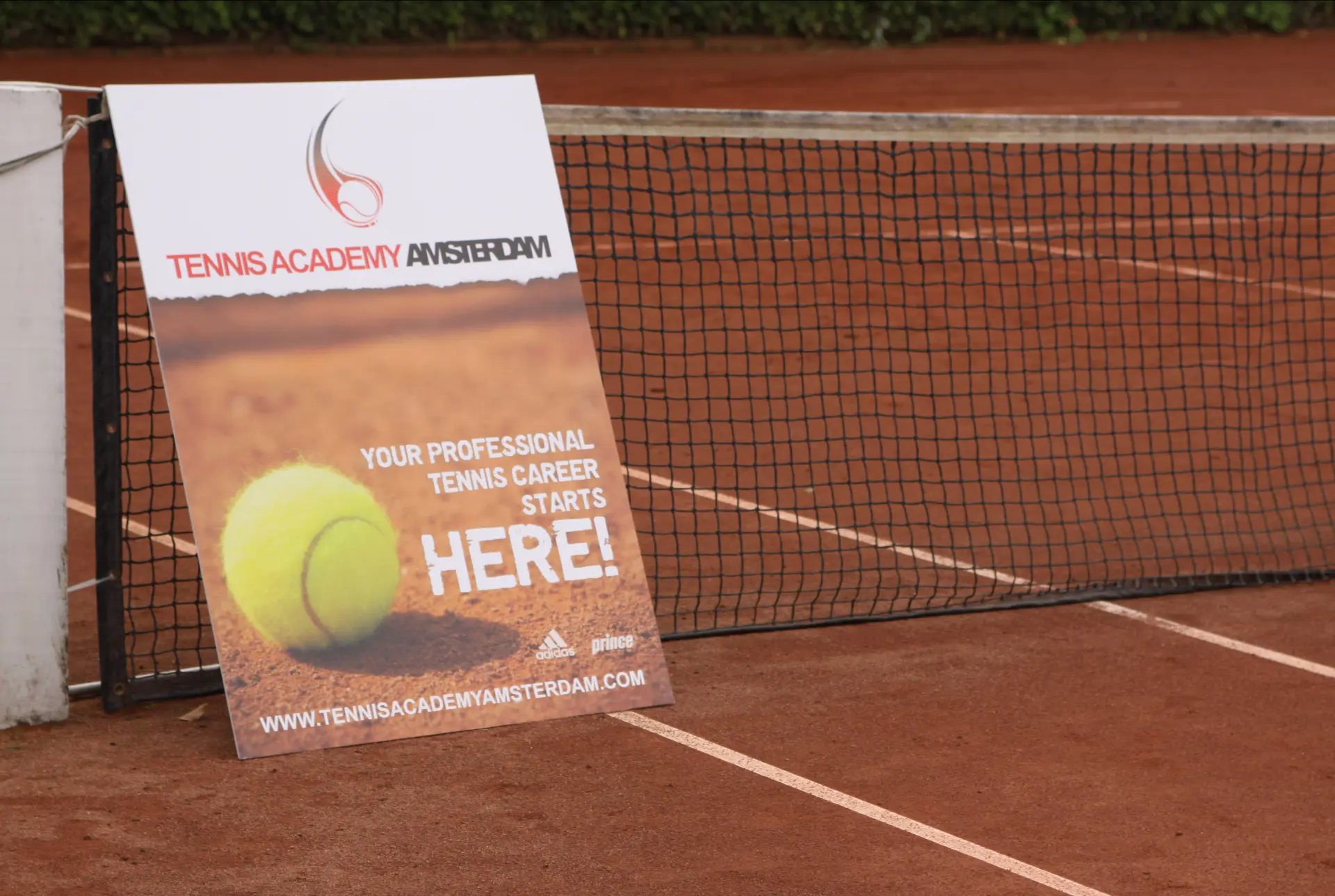 Baanposter van Tennis Academy Amsterdam naast het net op een gravelbaan, met de tekst “Your professional tennis career starts here” en het toenmalige webadres tennisacademyamsterdam.com.
