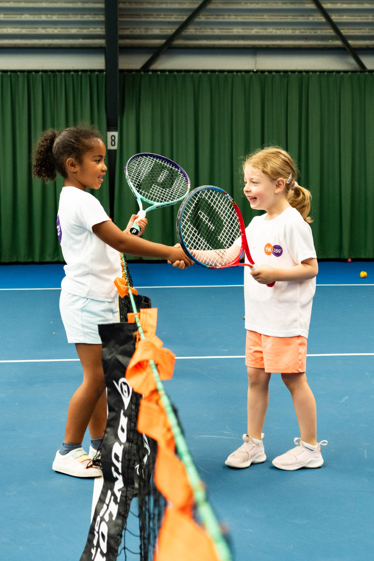 Twee tenniskids tikken hun rackets tegen elkaar bij het net tijdens indoor tennisles — 359.tennis