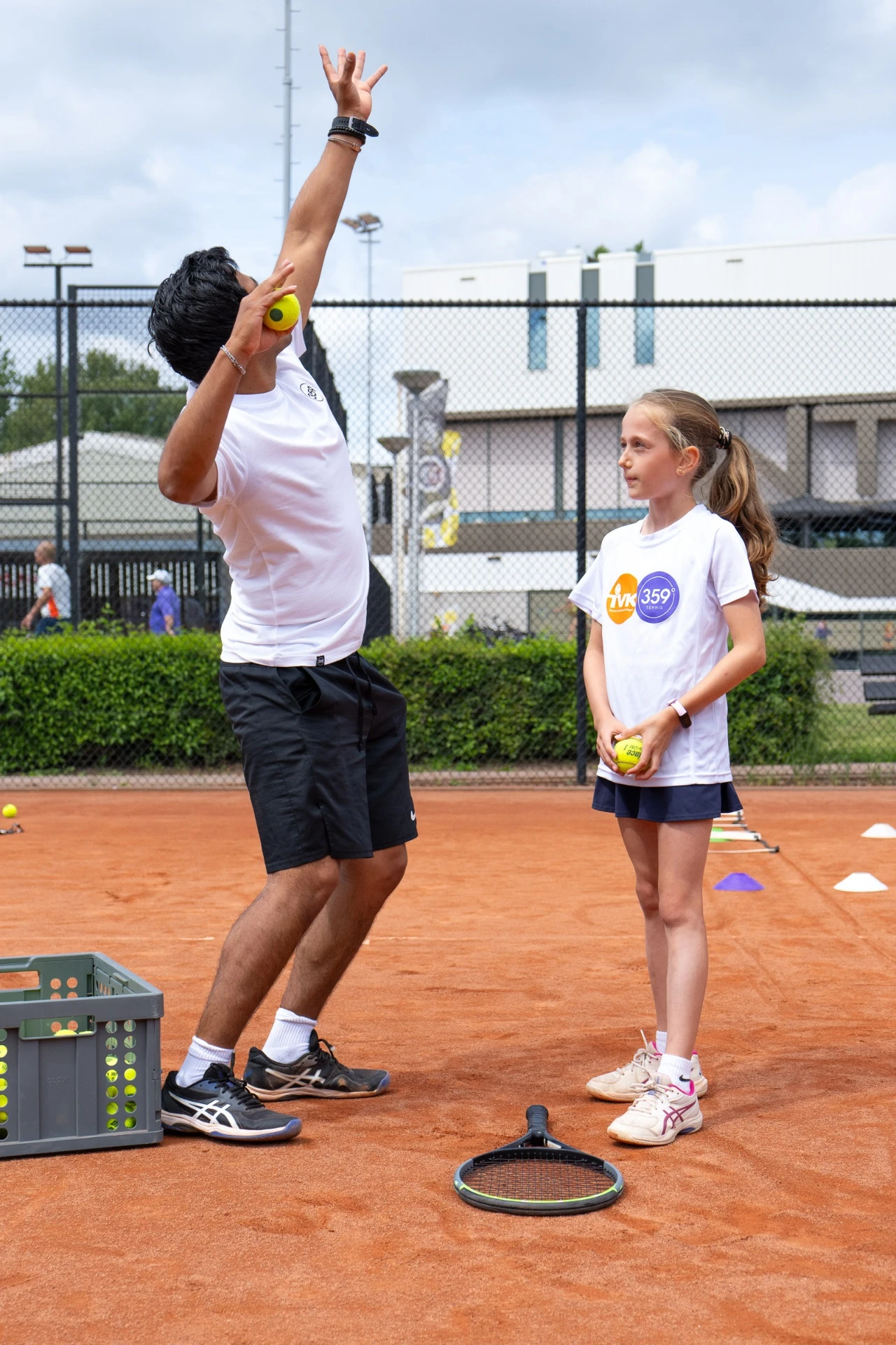 359-coach demonstreert de opslag aan een jong Tenniskids-lid op de gravelbaan